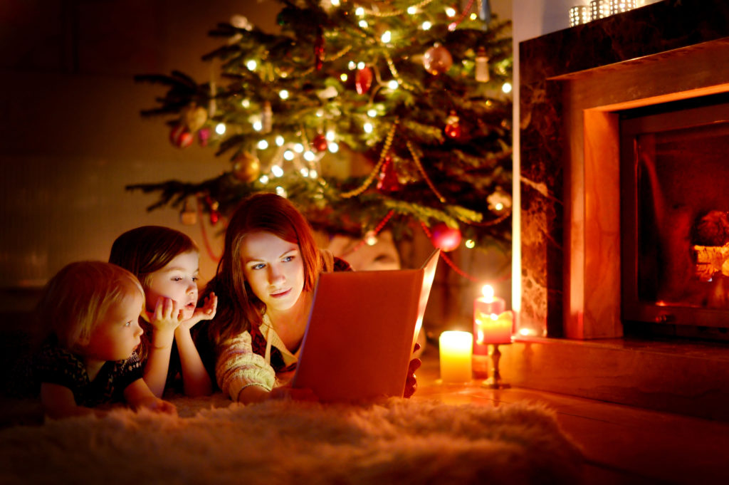 Young mother and her two little daughters reading a book by a Christmas tree in cozy living room in winter